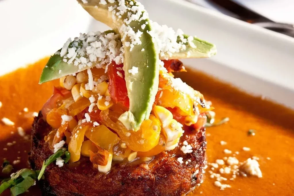 Seared patty topped with vegetables, avocado, cheese, and orange-red sauce.