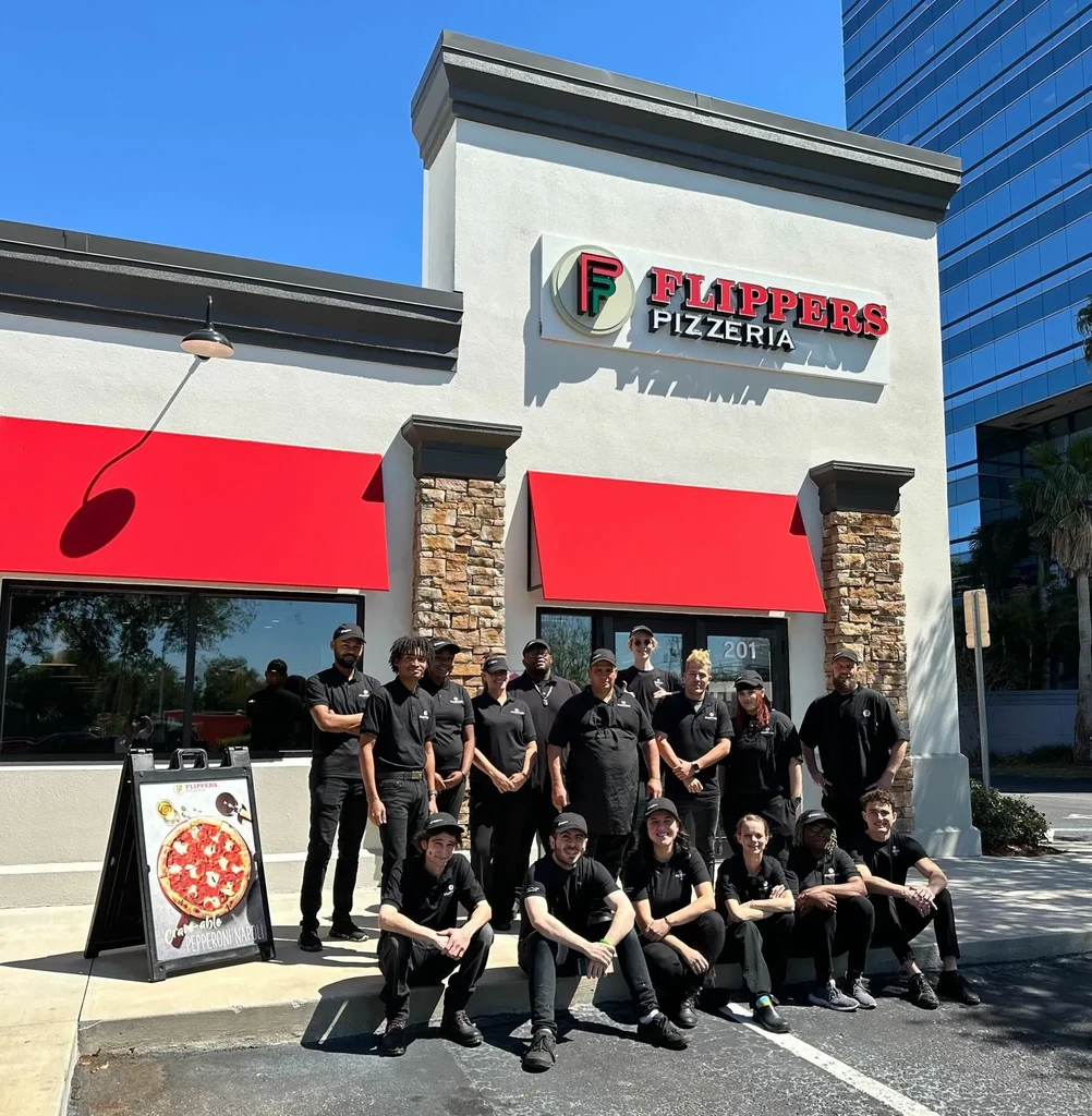 Sixteen staff members pose in front of Flipper's Pizzeria building.