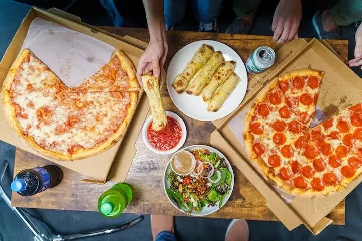 Overhead of hands sharing pizza, breadsticks, salad, and drinks.
