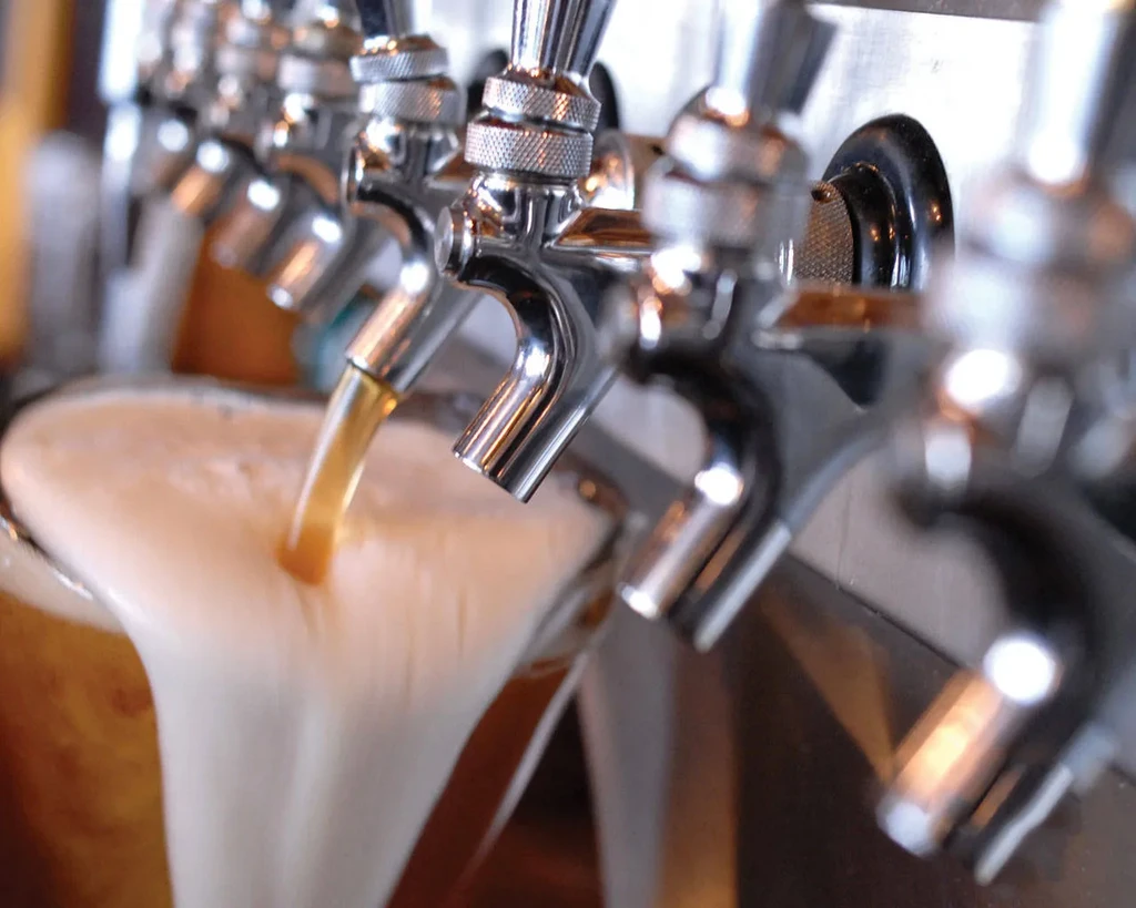 Close-up of metallic beer taps, one pouring amber beer with overflowing foam.
