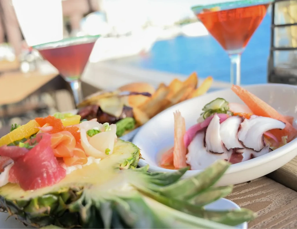 Outdoor spread of vibrant seafood, burger, fries, and drinks by water.