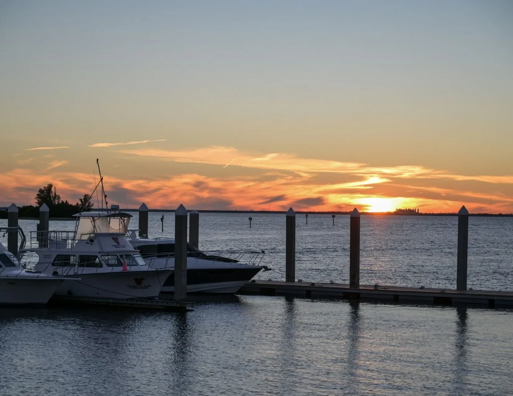Sunset over a marina with docked boats and a distant city.