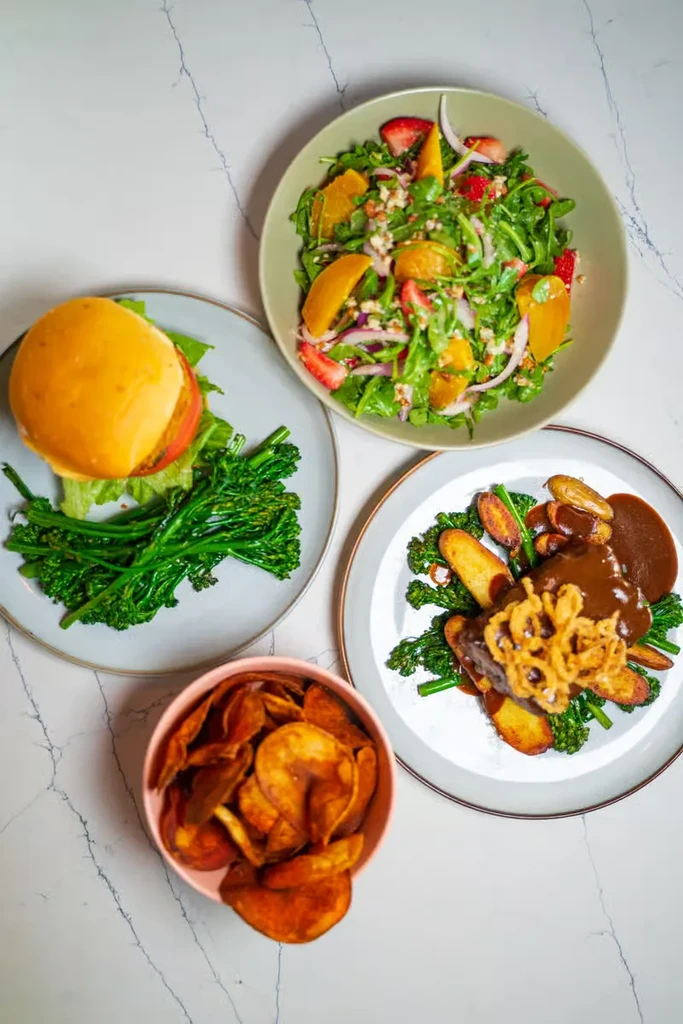 Four distinct meals: burger, salad, beef dish, and potato chips on marble.