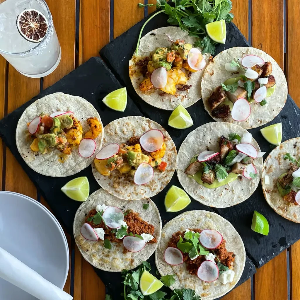 Overhead view of eight tacos with diverse fillings, lime wedges, and a margarita.