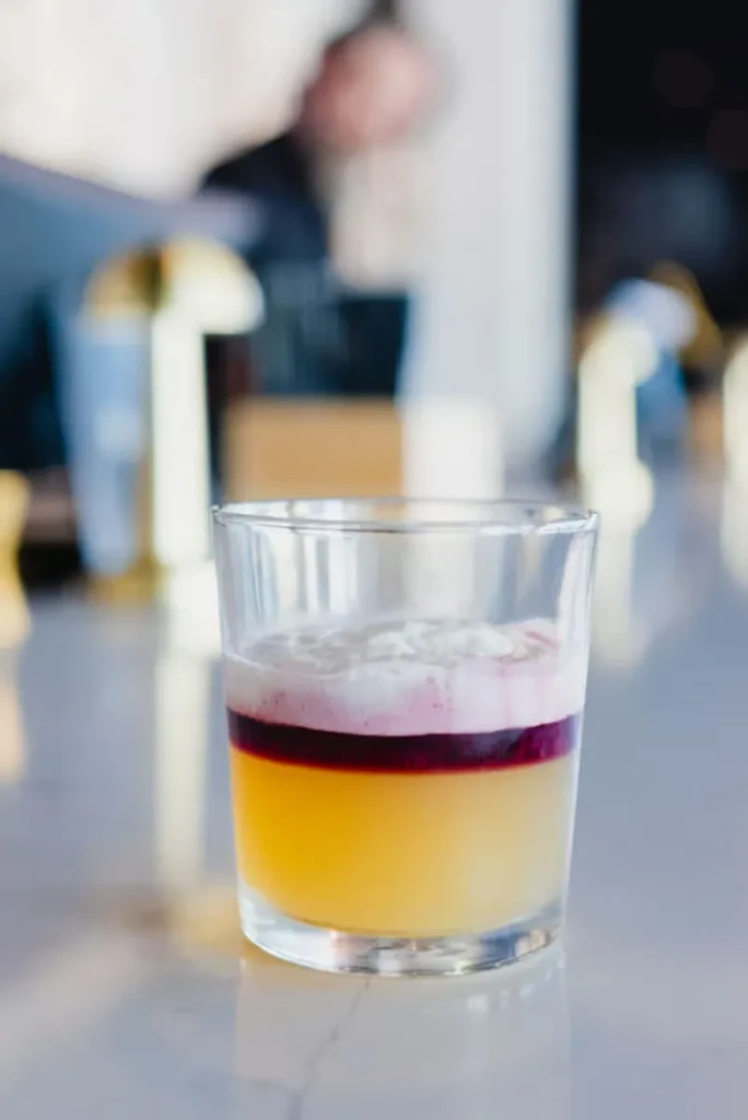 Close-up of layered drink: pale yellow, red band, frothy white top.