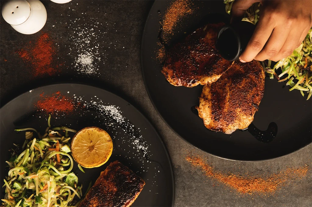 Top-down: hand pouring sauce over seasoned protein, green salad, and lemon.