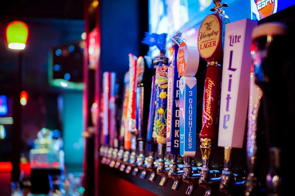 Colorful, branded beer tap handles in focus at a blurred, lively bar.