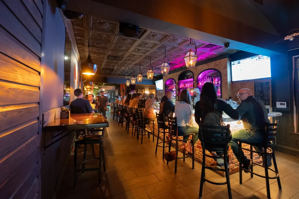 Dimly lit bar with people, ornate ceiling, and amber-magenta lights.