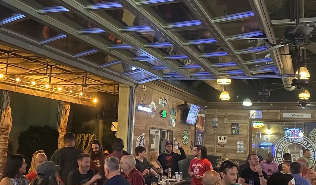 Lively indoor-outdoor bar with glass roof, illuminated by string lights and neon.