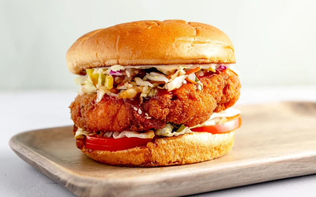 Crispy fried chicken sandwich with coleslaw, tomato, and lettuce on wooden board.