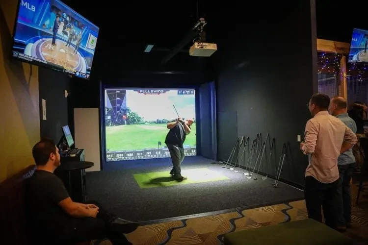 Inside a modern venue: man swings golf in simulator, MLB on TV.