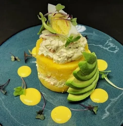 Layered yellow potato dish, filling, avocado, egg, microgreens, and sauce.