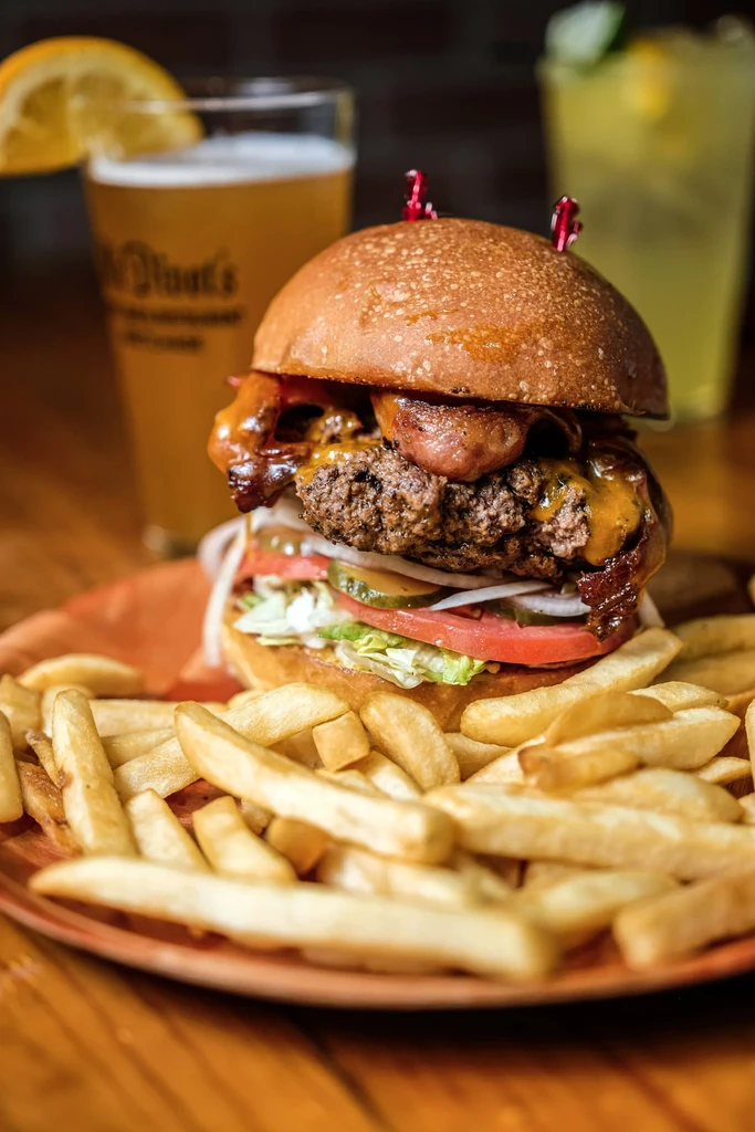 High-stacked burger, golden fries, and blurred drinks on an orange plate.