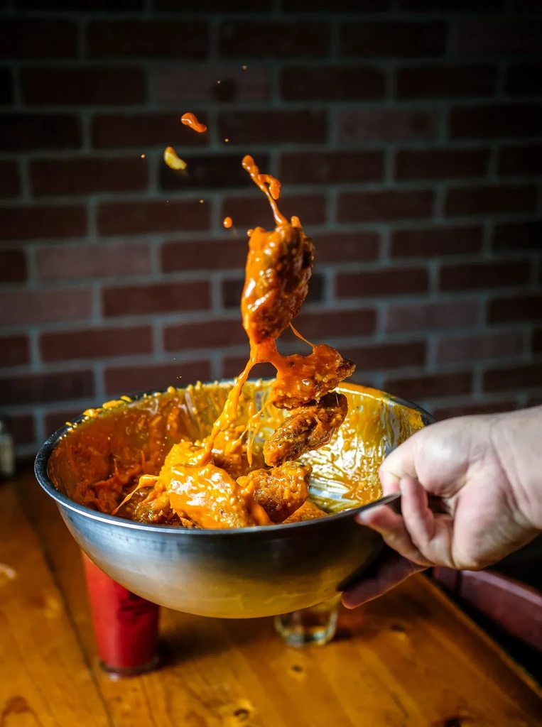 Hand tossing sau saucy chicken wings in a metal bowl.