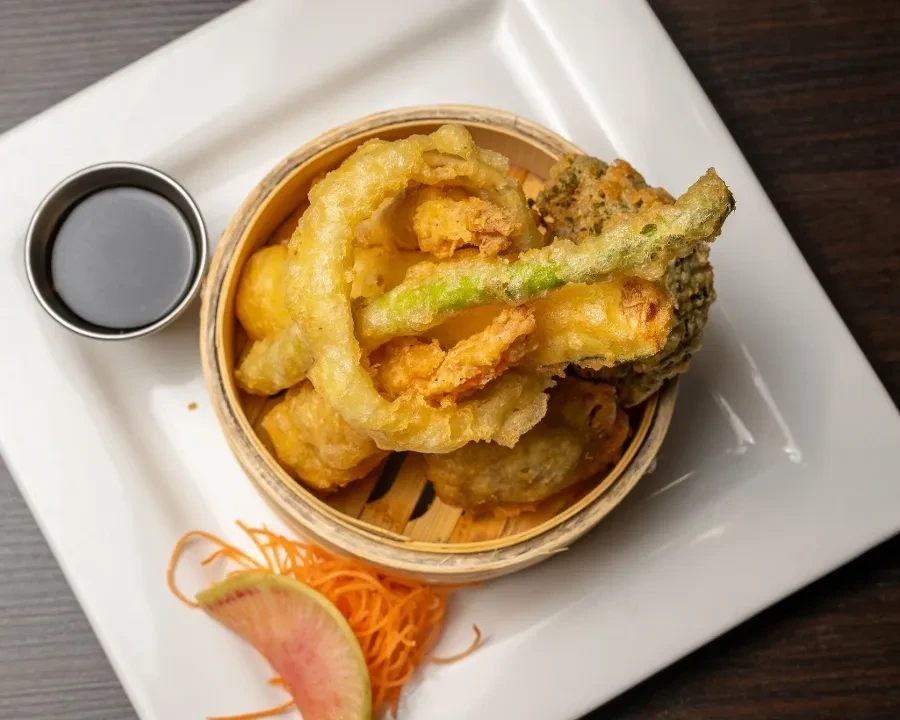 Golden-brown tempura vegetables in a steamer basket with dipping sauce.