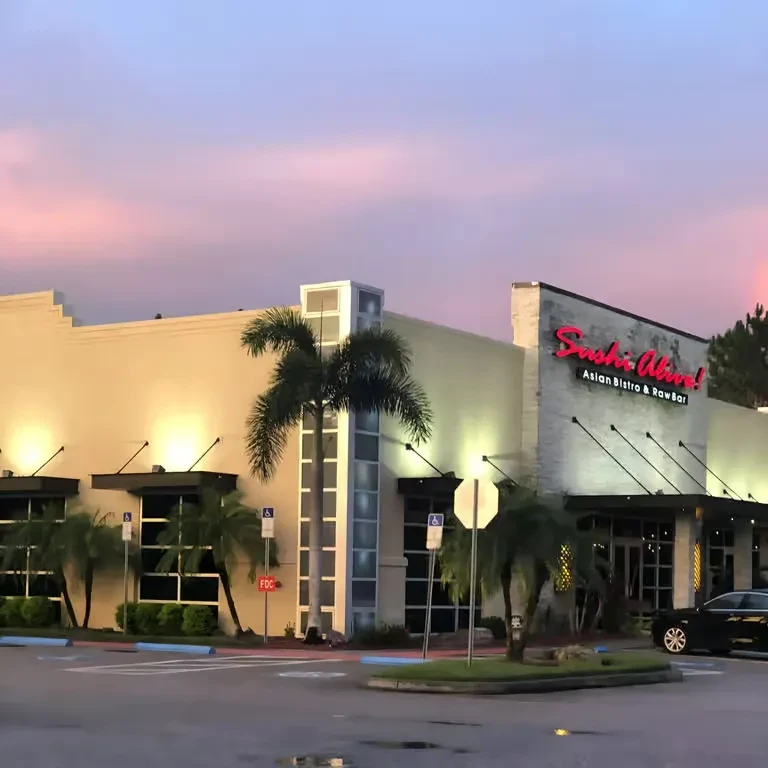 "Sushi Alive!" restaurant exterior at dusk, pink neon sign, palm trees.