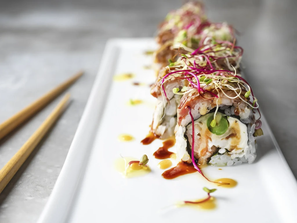 Sushi rolls with colorful sprouts and drizzled sauces on a white plate.
