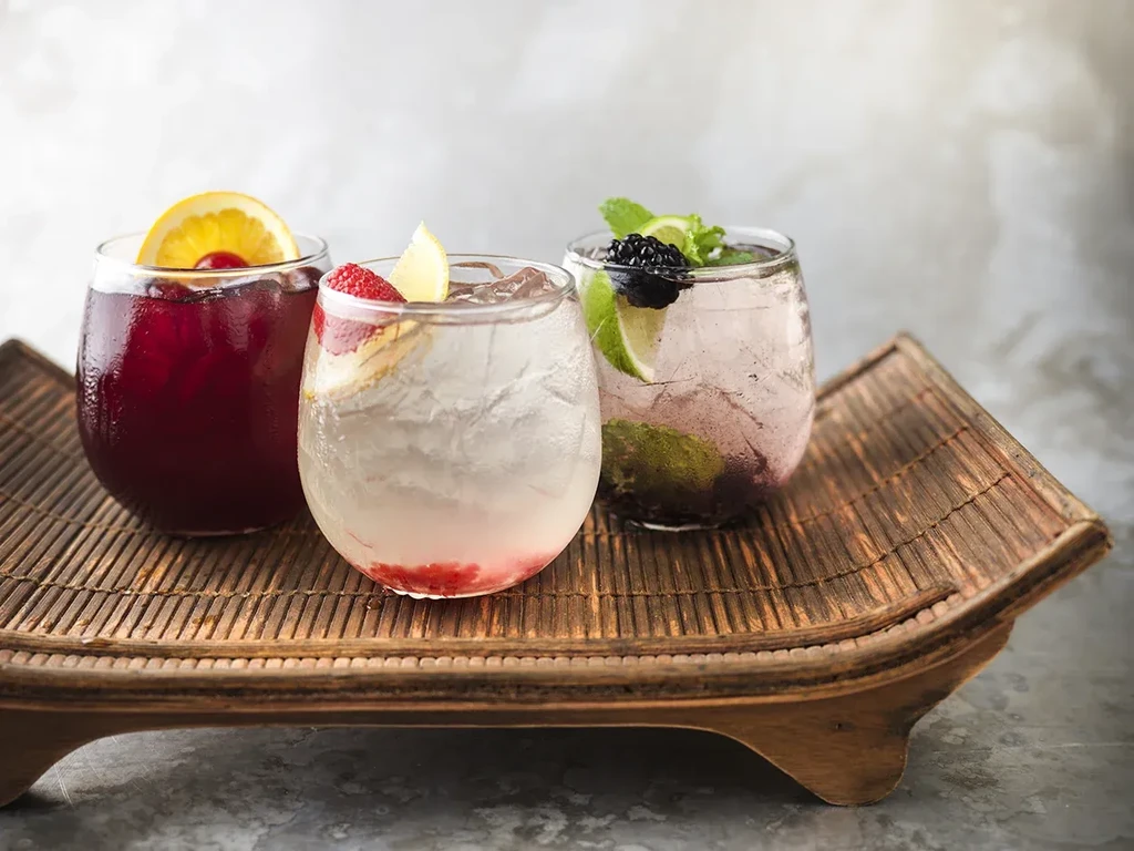 Three colorful beverages on a wooden tray with fruit garnishes.