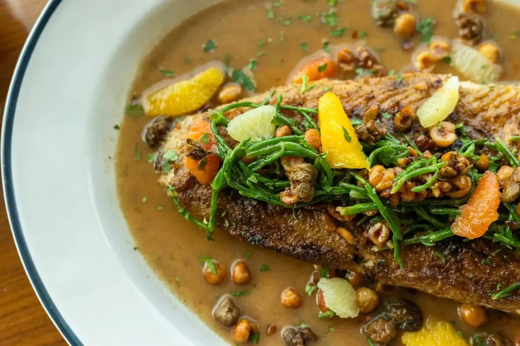 Close-up of pan-seared fish, green garnish, nuts, citrus, and sauce.
