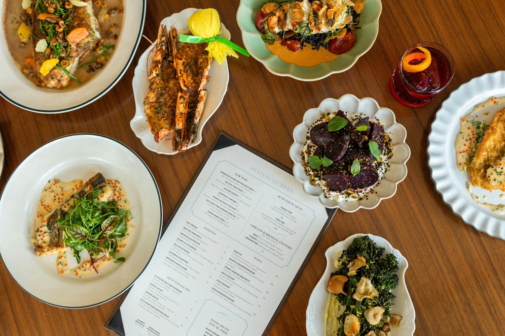 Overhead view of dining table with diverse plated seafood dishes, cocktail, and menu.