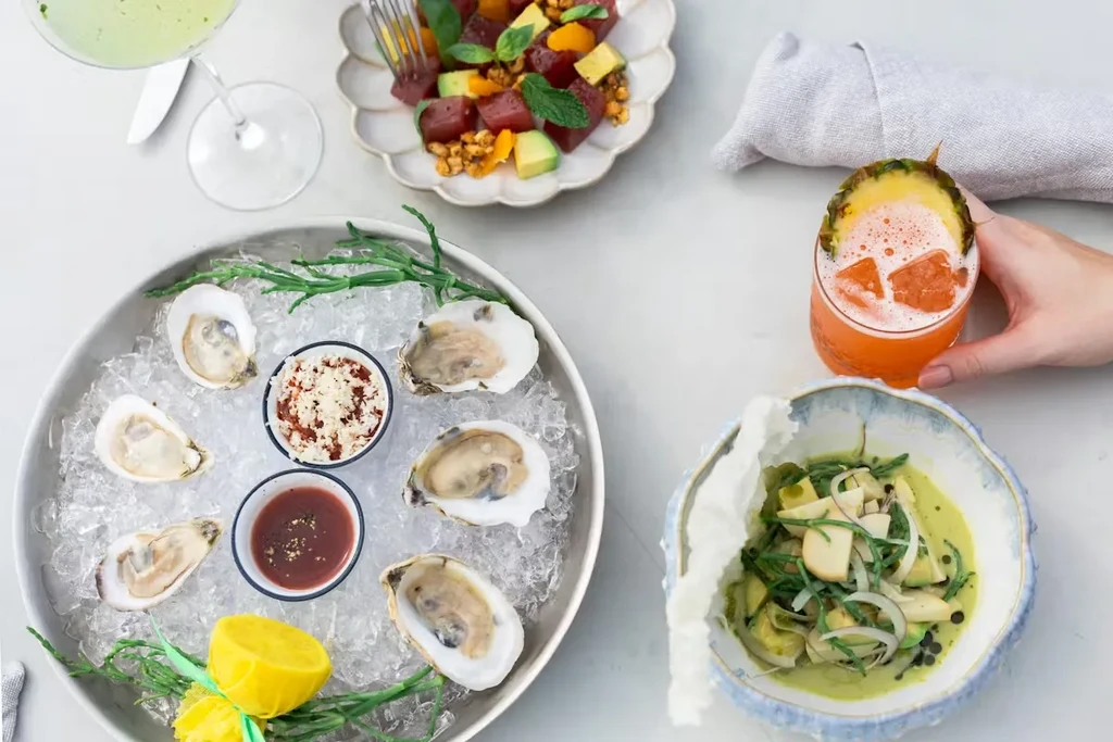Flat lay of gourmet oysters, poke, green dish, and cocktails.