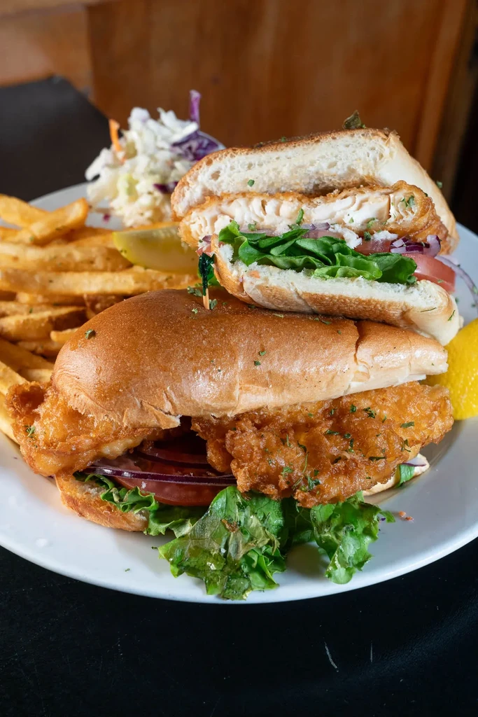 Plate of fried fish sandwich, french fries, coleslaw, pickle, and lemon.