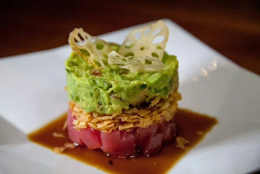 Plated tower of tuna, guacamole, crispy flakes, crisps, and sauce.