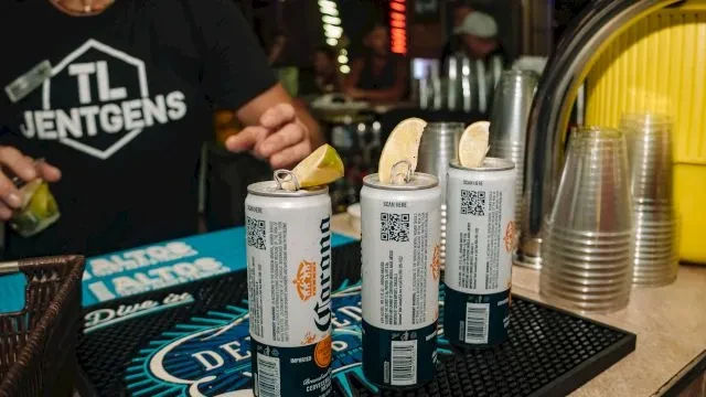 Three open Corona beers with lime wedges; a person squeezes lime.