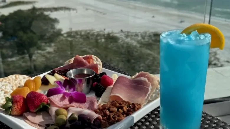 Colorful charcuterie platter and blue cocktail with a beach background.