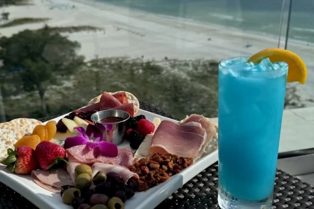 Charcuterie board and blue cocktail overlooking a sandy beach.