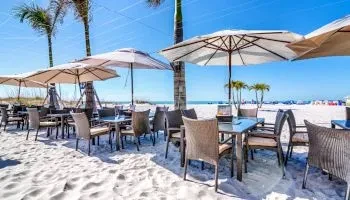Beach dining area with tables, chairs, umbrellas, palm trees, and ocean.