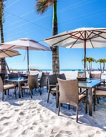 White sand beach restaurant with empty tables, umbrellas, and palm trees.