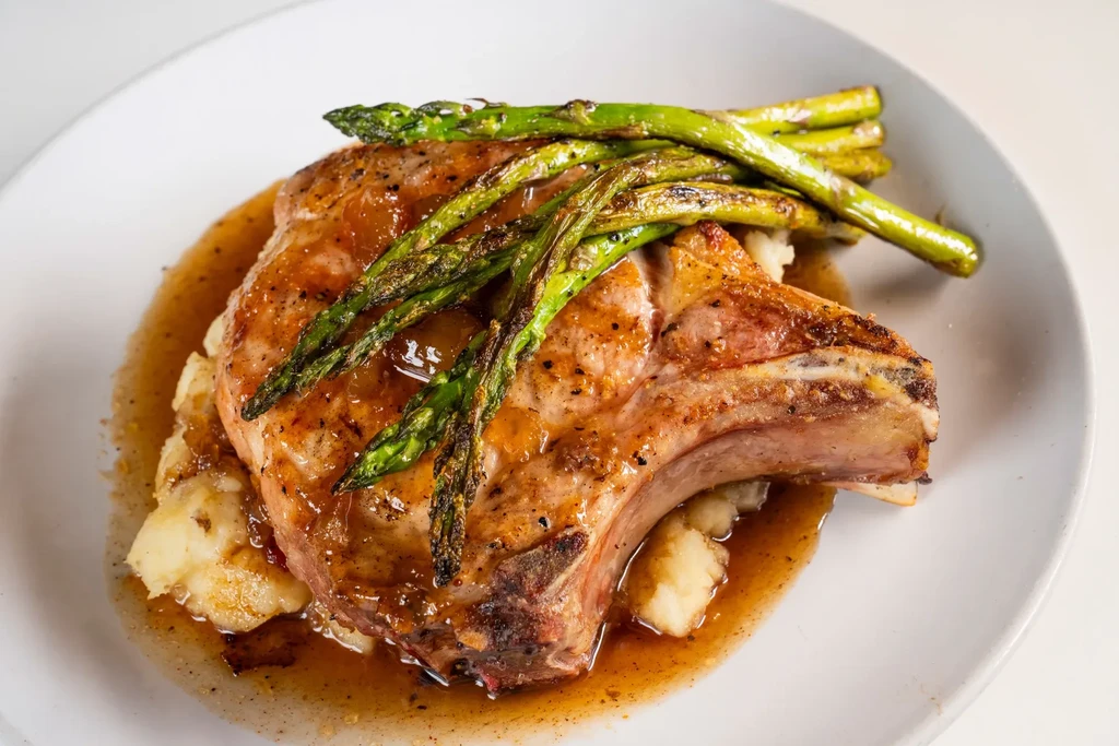 Plated seared pork chop, mashed potatoes, and roasted asparagus with sauce.