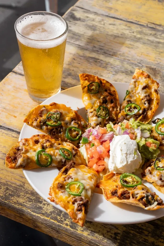 Overhead: Cheesy mini-tacos with toppings and beer on a rustic table.