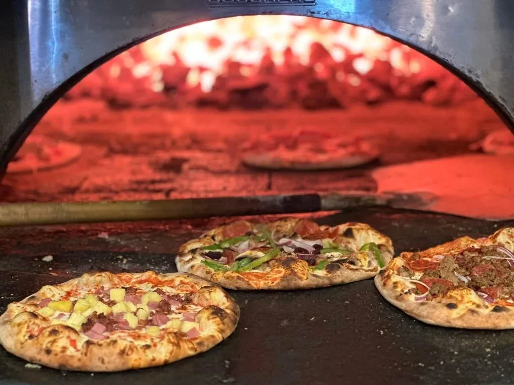 Three artisanal pizzas cooking in a glowing wood-fired oven.