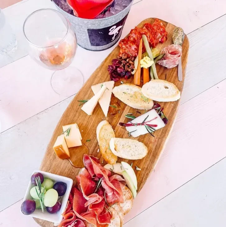 Surfboard charcuterie board with cheeses, meats, fruit, and rosé wine.