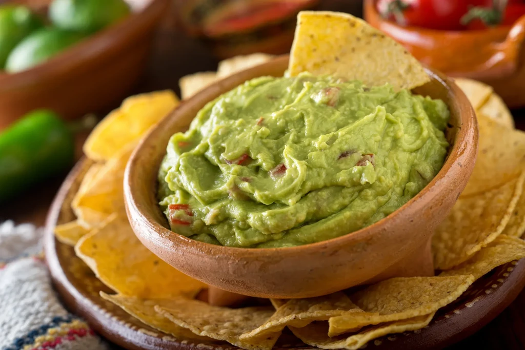 Terracotta bowl of chunky guacamole and tortilla chips, one inserted.