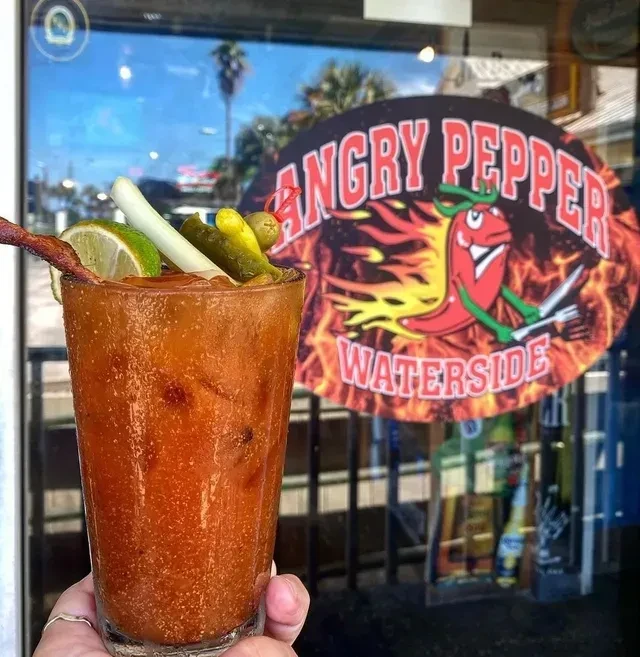 Hand holds heavily garnished Bloody Mary; blurred "Angry Pepper" sign.