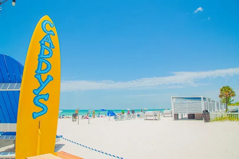 Yellow Caddy's surfboard on sunny beach with ocean, people, cabana.