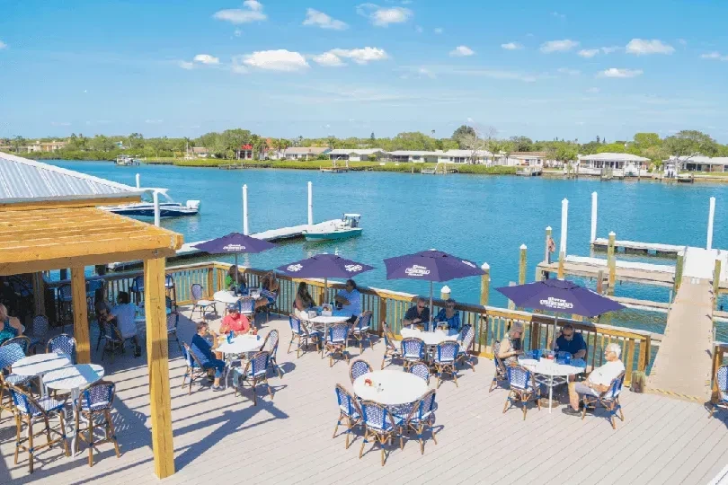 Vibrant outdoor restaurant patio with diners and Corona umbrellas by calm water.