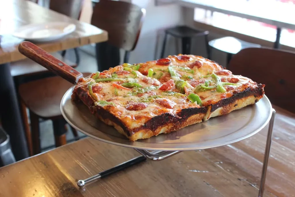 Square-cut pizza with cheese, pepperoni, green peppers on a wooden table.