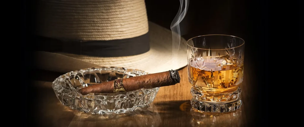 Dramatic still life with lit cigar, whiskey glass, and fedora hat.