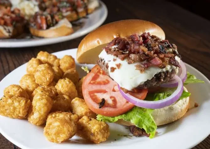Open-faced burger with bacon, cheese, vegetables, and golden tater tots.