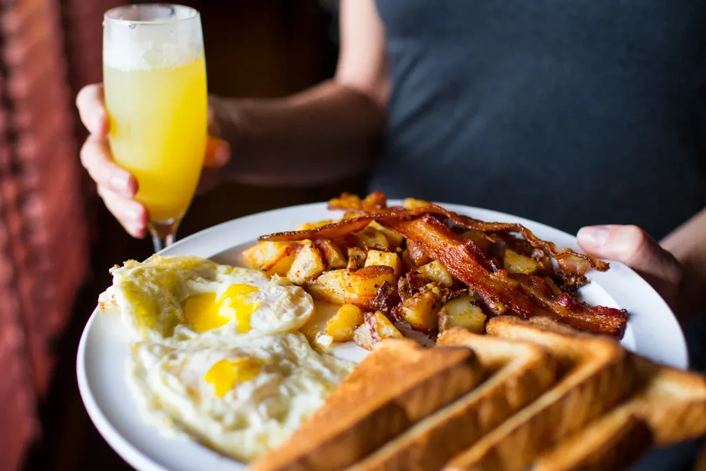Person holding a plate of fried eggs, bacon, potatoes, and a drink.