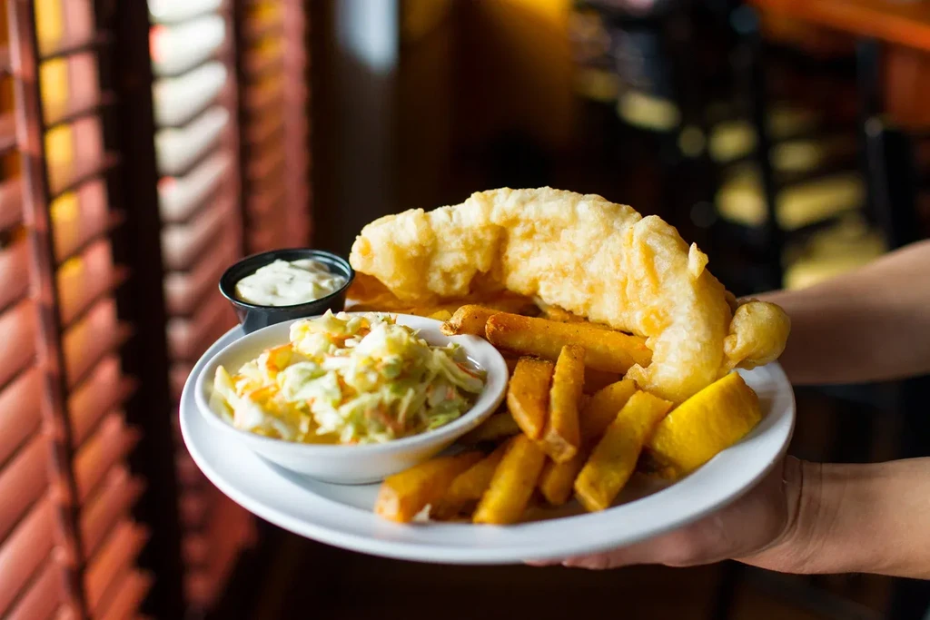 Hands hold a plate of battered fish, chips, coleslaw, and sauce.