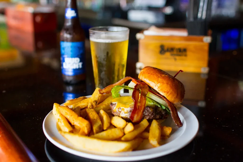 Burger, fries, Bud Light bottle, and beer on a dark bar surface.