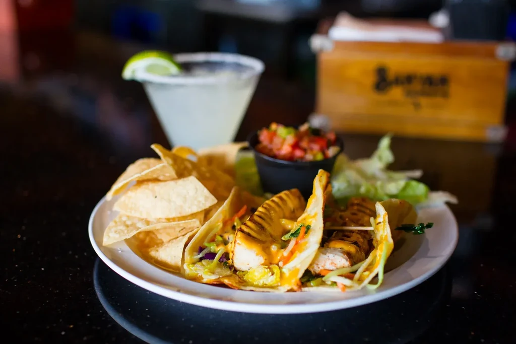 Three tacos, tortilla chips, salsa, and a margarita on a dark table.