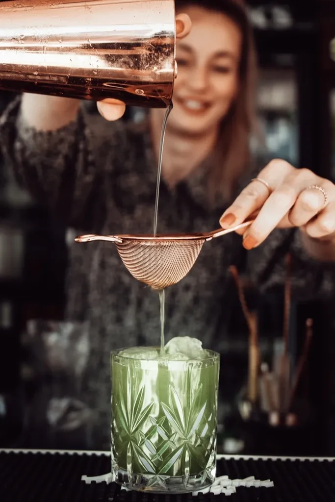 Hand straining green cocktail into crystal glass, blurred bartender.