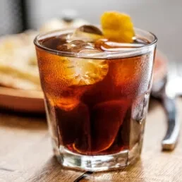 Amber drink with ice and citrus peel in a faceted rock glass.