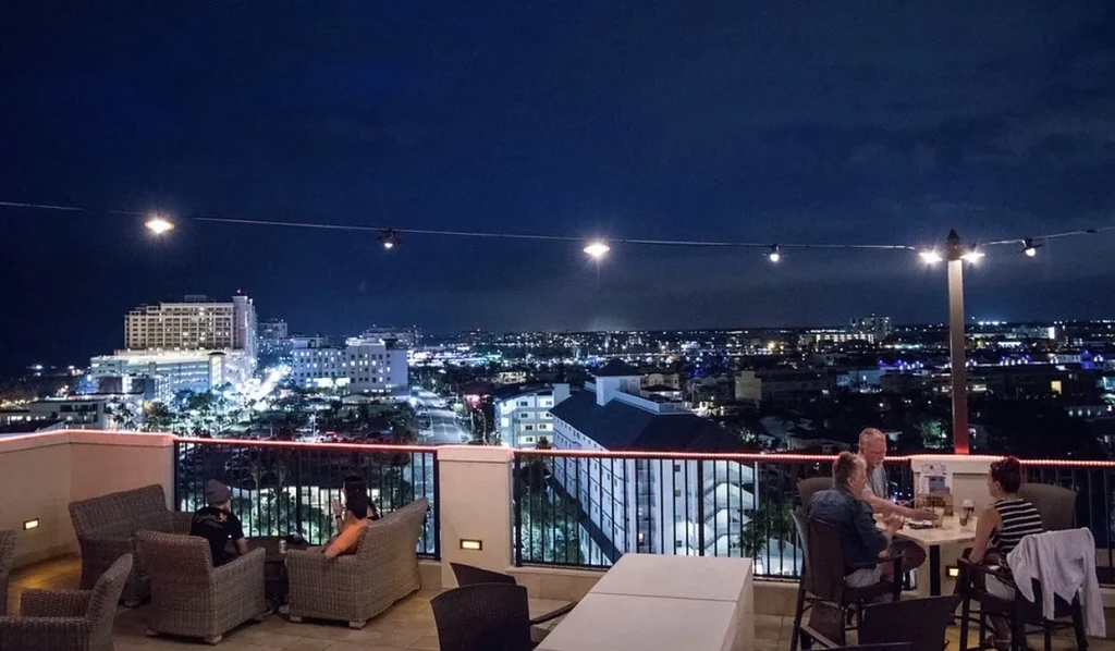Nighttime rooftop bar with seated patrons and city skyline view.